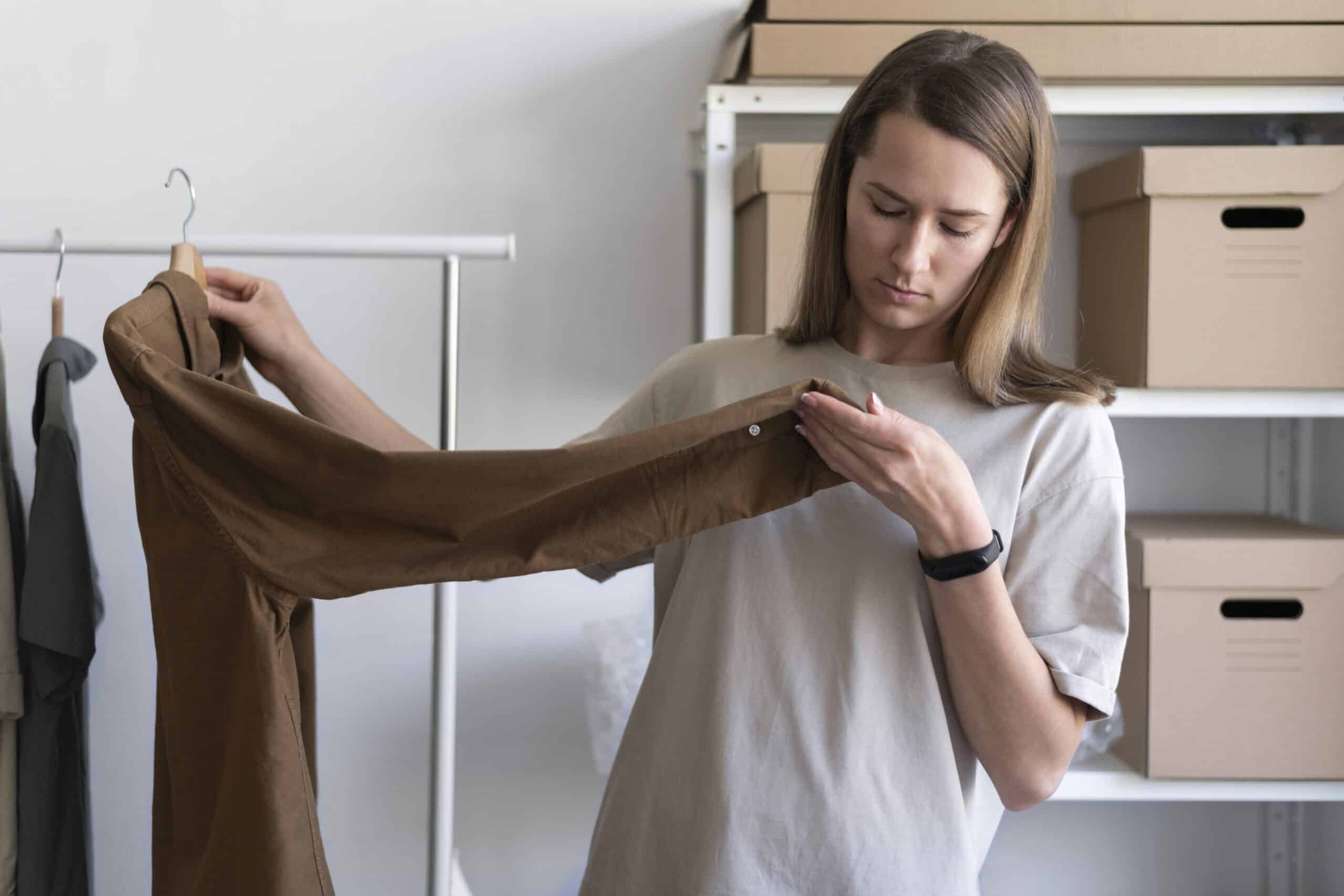 A woman closely inspecting the sleeve of a brown shirt in a storage room.